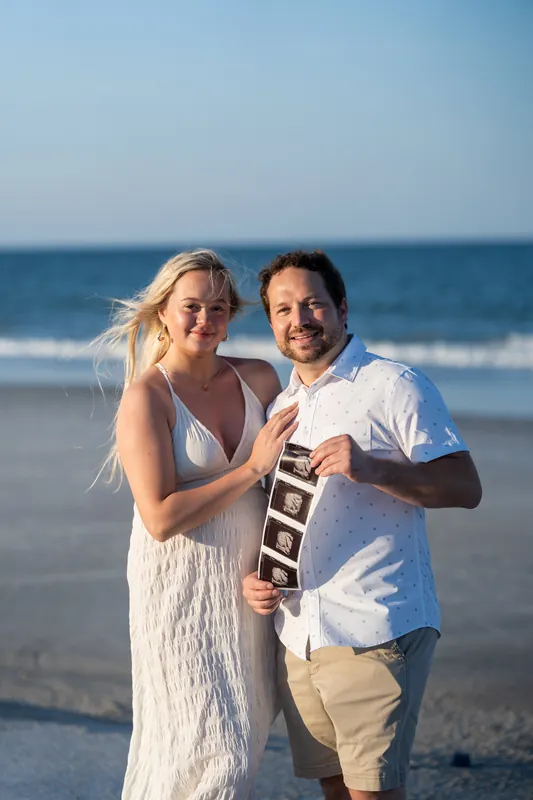 Paige and Steve pose together on the beach holding up a ring box and smiling at the camera with the ocean behind them.