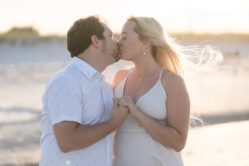 Steve kisses Paige's cheek at golden hour on Vilano Beach, her blonde hair blowing in the breeze as she smiles.