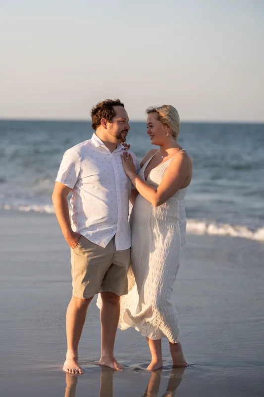 Steve in a white button-up shirt stands facing Paige in a cream linen dress as she adjusts his collar on the beach.