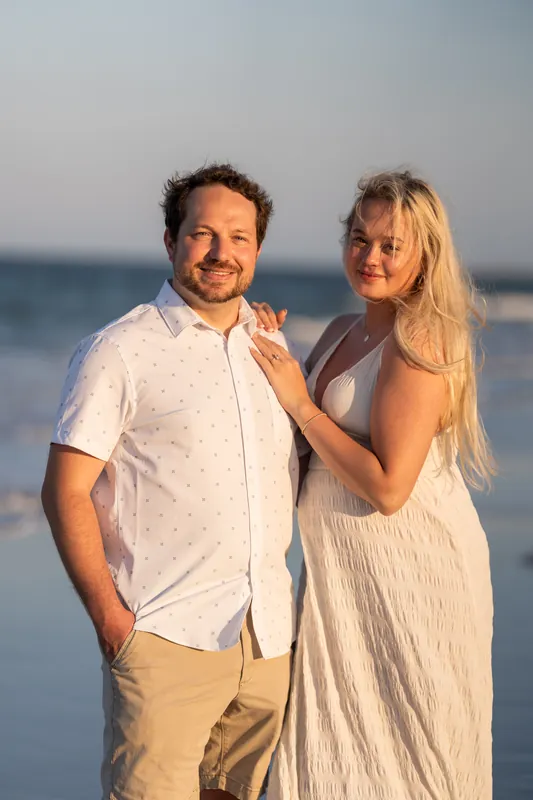 Steve and Paige pose together on the sand with the ocean and sky behind them, both smiling at the camera.