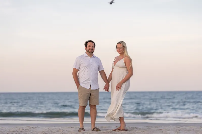 Steve and Paige hold hands while standing on the wet sand at the ocean's edge, the horizon stretching behind them.