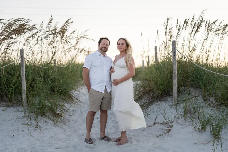 Steve and Paige stand between dune grasses on the beach, Paige in white and Steve in a light shirt.