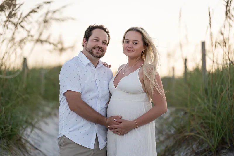 Steve places his hand on Paige's belly as they smile at the camera, tall beach grasses framing them on either side.