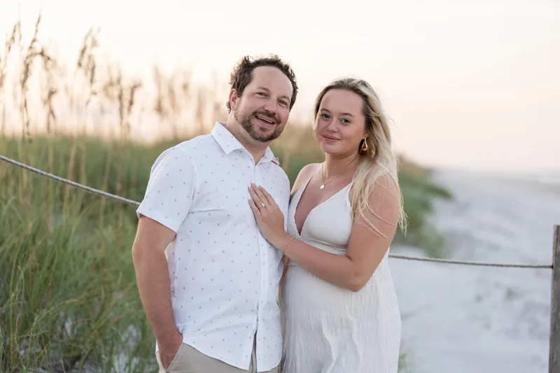 Steve and Paige stand together among beach grasses at sunset, smiling as Steve holds Paige close to his chest.