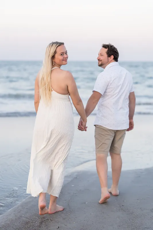 Paige and Steve walk hand-in-hand along the shoreline at dusk, both barefoot in the wet sand.