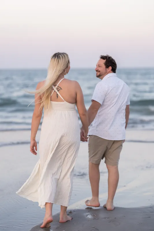 Paige looks back at Steve while they hold hands wading through shallow water as the sun sets over the ocean.
