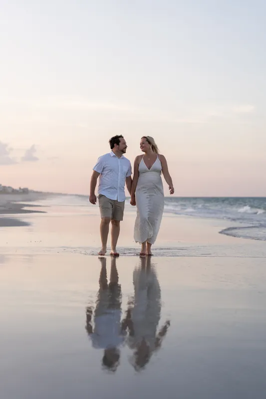 The couple walks toward the camera across wet sand reflecting the twilight sky at the water's edge.