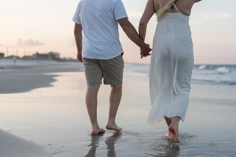 Steve and Paige stroll through shallow surf holding hands, with golden hour light illuminating the beach behind them.