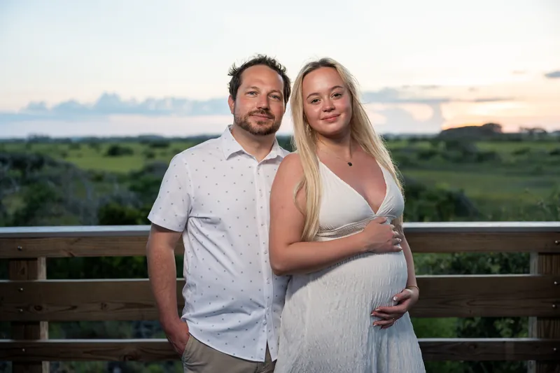 Steve and Paige stand on a wooden railing at Vilano Beach at golden hour, overlooking marshland as she cradles her belly.