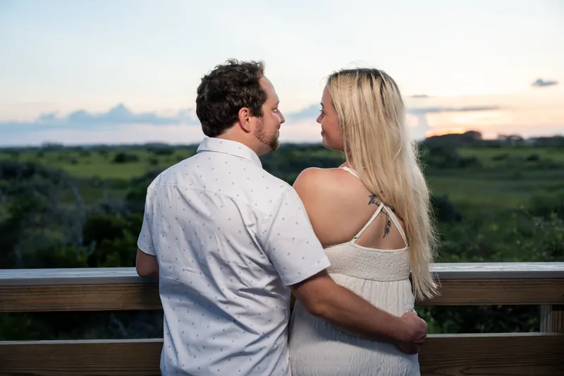 Steve kisses Paige's cheek as they embrace at the railing with the beach landscape and evening sky behind them.