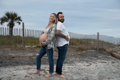 Fun maternity portrait of couple standing back-to-back showing off the baby bump, with beach dunes and palm trees behind them.