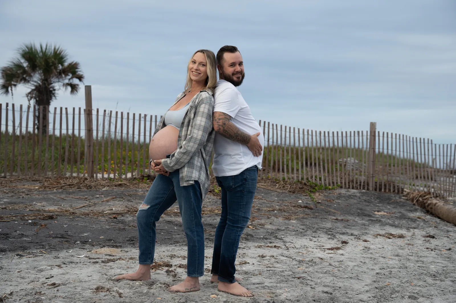 Fun maternity portrait of couple standing back-to-back showing off the baby bump, with beach dunes and palm trees behind them.