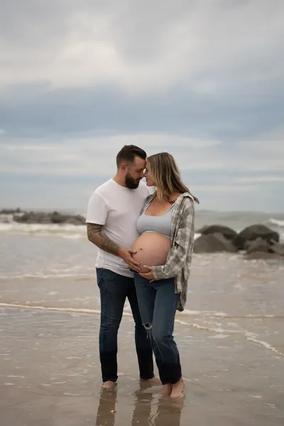 Intimate moment as couple stands foreheads together in the shallow water, hands cradling the baby bump with jetty rocks in the background.