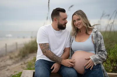 Couple sits on a wooden bench among the sea oats at Vilano Beach, Taylor looking adoringly at Stormi as she smiles at the camera.