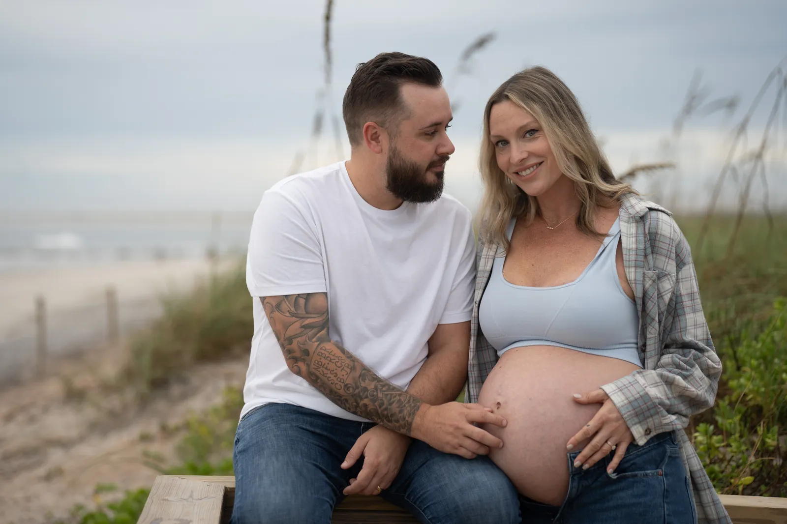 Couple sits on a wooden bench among the sea oats at Vilano Beach, Taylor looking adoringly at Stormi as she smiles at the camera.