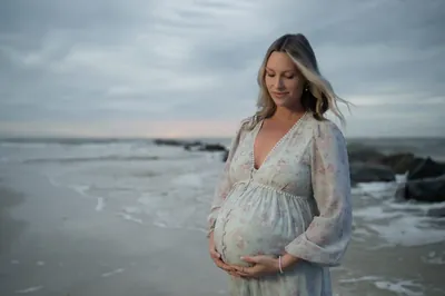 Expectant mother in floral dress looks down at her baby bump with a peaceful expression, ocean and jetty rocks behind her.