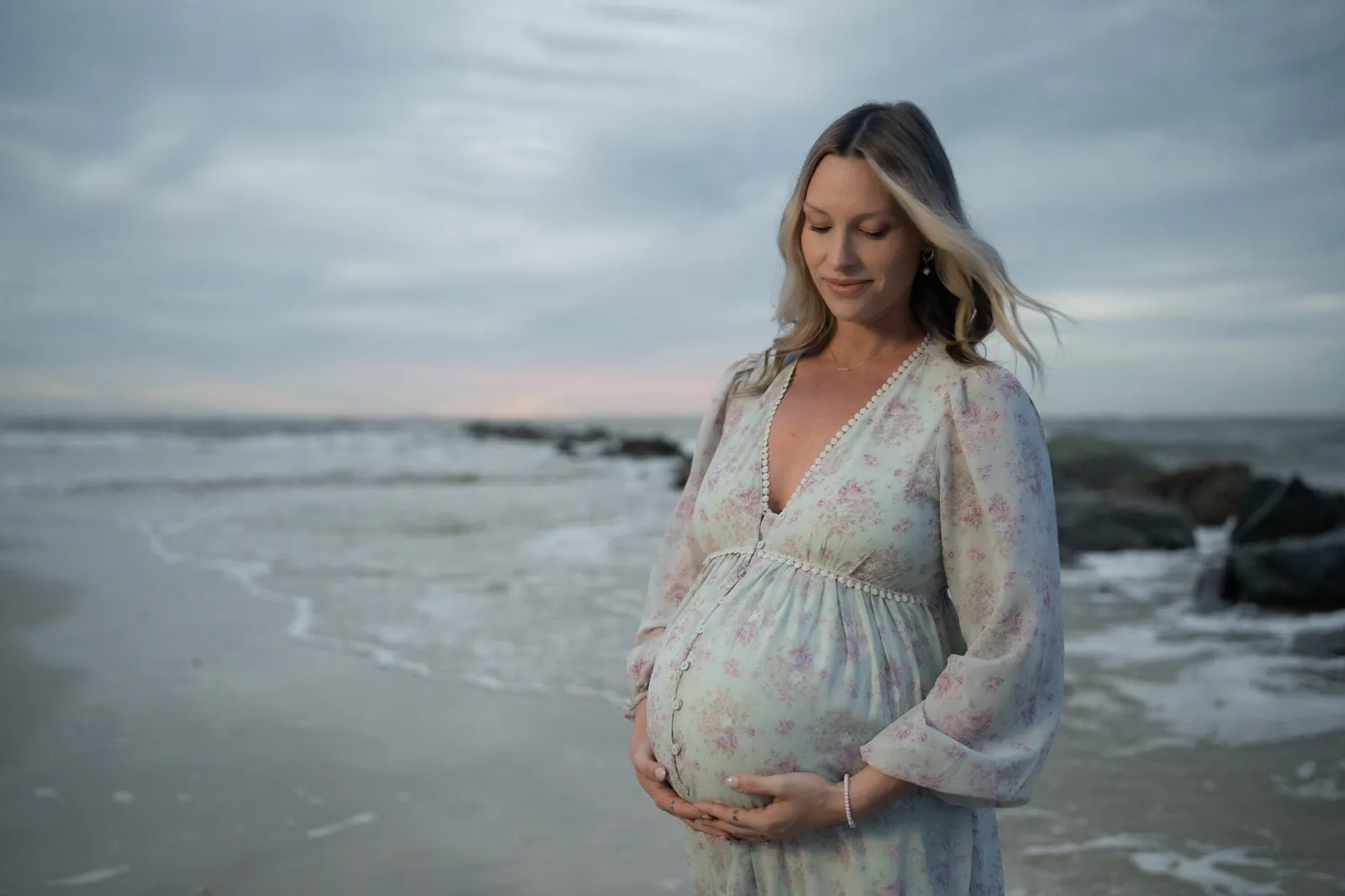 Expectant mother in floral dress looks down at her baby bump with a peaceful expression, ocean and jetty rocks behind her.