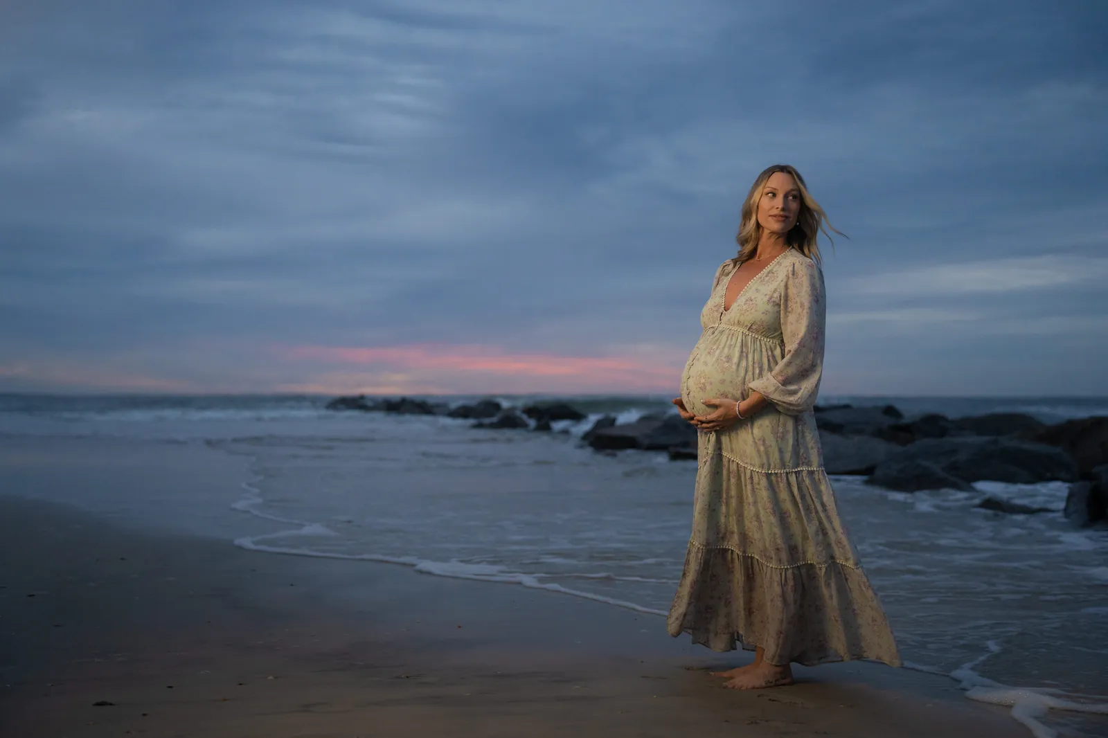 Expectant mother in flowing floral dress stands on Vilano Beach at sunrise, cradling her baby bump with pink and blue sky behind her and jetty rocks in the distance.