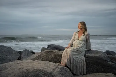 Pregnant woman sits gracefully on jetty rocks at Vilano Beach, looking out at the ocean in her flowing floral dress.