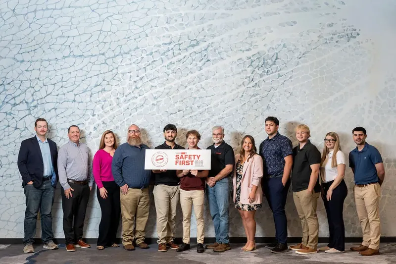 A group of twelve people in business casual attire stands in a line holding a Superior Party check against a textured white