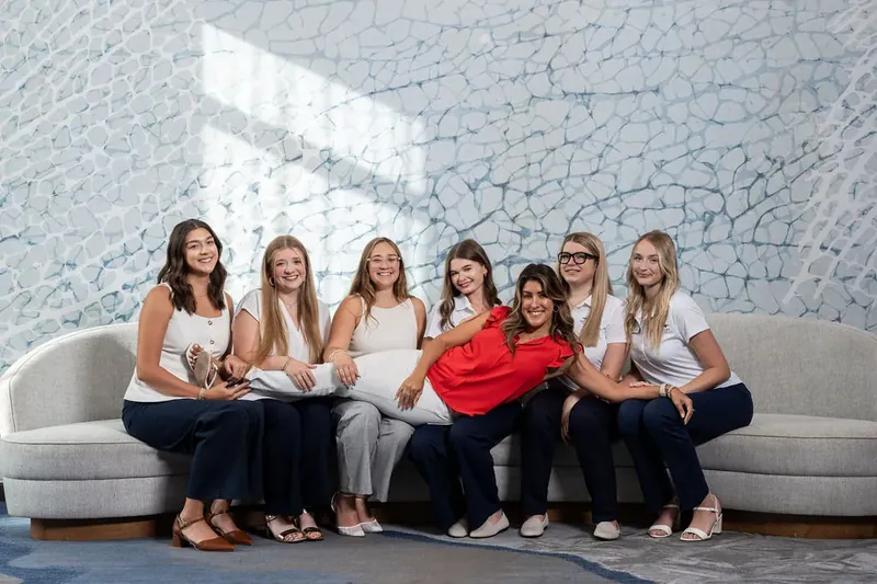 Seven women sit on a curved gray sofa, with one wearing a bright red top among neutral-toned outfits, against a textured wall