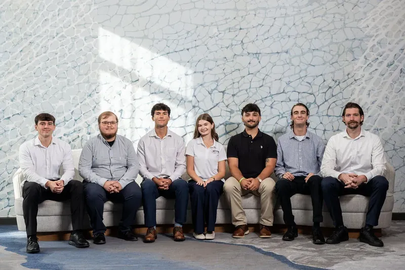 Seven men sit in a row on a gray bench against a crackle-textured wall, wearing casual button-ups and neutral tones