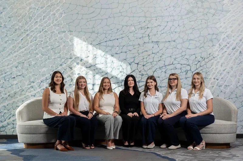 Eight women sit on a curved gray sofa in white and black tops, arranged against a textured wall with one figure in all black