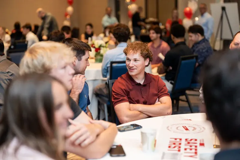 Person in rust-colored shirt smiles while conversing at a table during an indoor corporate event with attendees and red