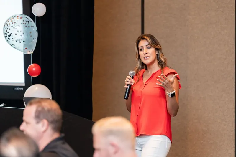 Woman in bright red button-up shirt holds a microphone while speaking to an audience in an indoor event space.