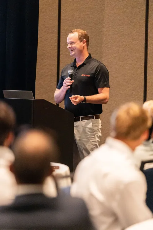 Man in black polo shirt addresses seated attendees while holding a microphone at a corporate gathering.