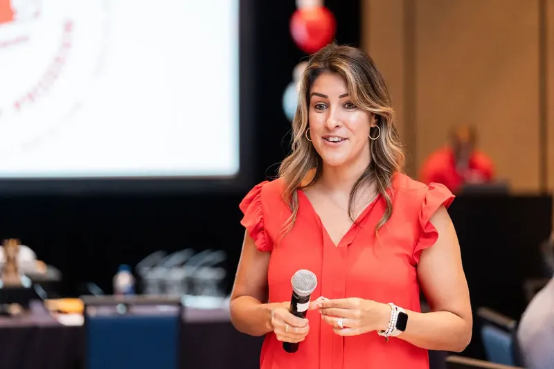 Woman in coral ruffled top holds a microphone and smiles while speaking to an audience at an indoor event.
