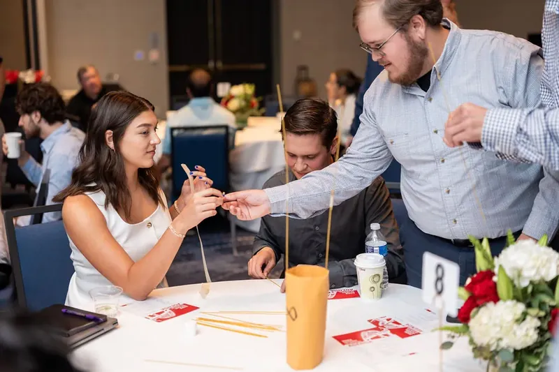 Two people at a table reach toward a game piece while others participate in an interactive team activity behind them.