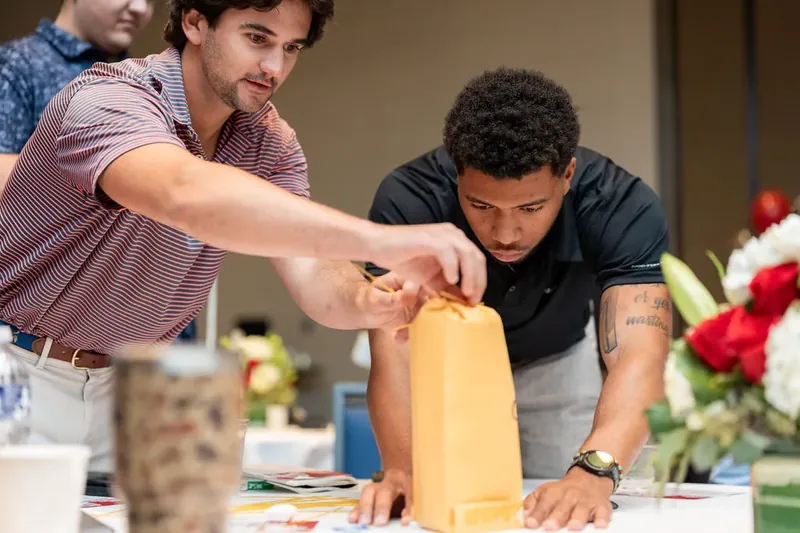 Two men lean over a yellow structure at a table, working together to build or adjust it during a team competition.