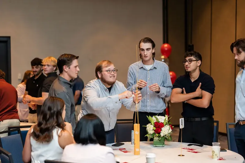 Mixed group of employees in casual attire gather around a table with wooden crafts and red flowers, engaged in conversation