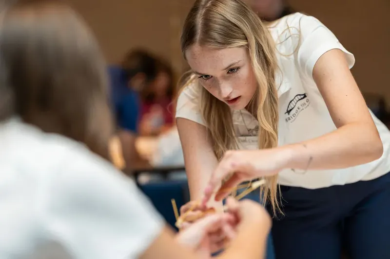 Staff member in white shirt leans forward to assist a participant's hands with a string-based puzzle or craft activity.