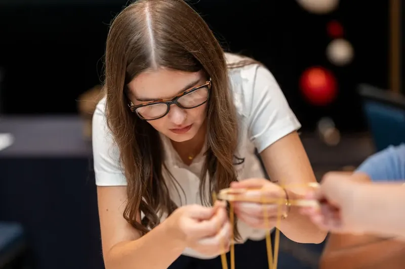 Person wearing glasses focuses intently while holding golden twine connected to a wooden structure on the table in front