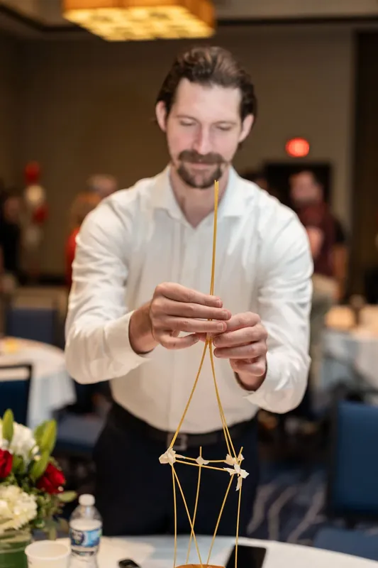 Man in cream shirt extends his hands to show a completed geometric wooden structure made with golden string at an indoor