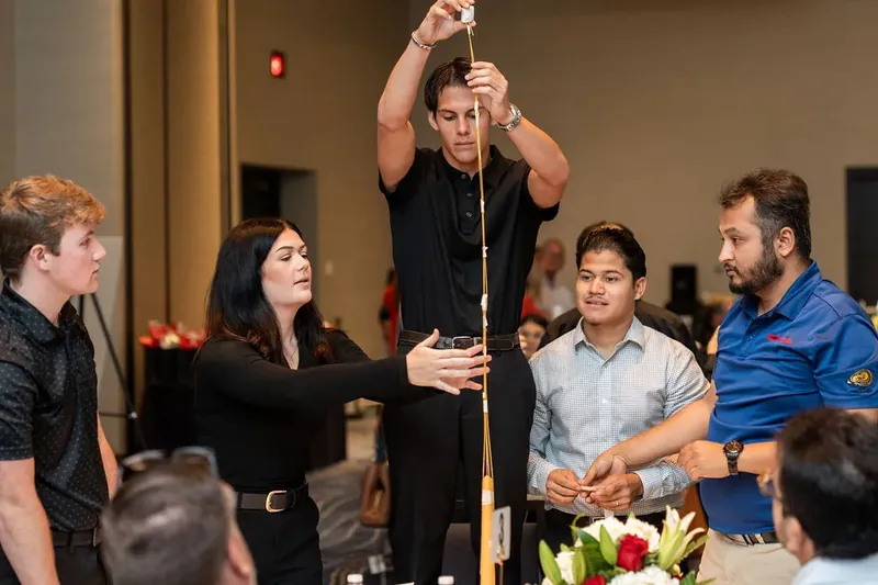 Team members gather around a tall wooden structure during an interactive game, with a man in black holding the top piece