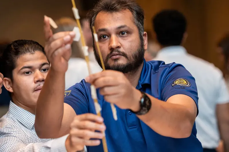 Man in a blue polo shirt concentrates on placing a wooden block onto a tall tower while a young participant watches beside