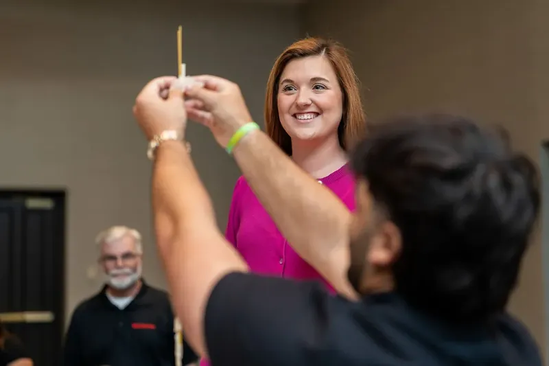 Woman in a bright pink top smiles while carefully positioning a wooden block on the tower structure during the activity.
