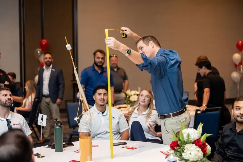 Participant in a denim shirt reaches up to place a block on the towering wooden structure as seated colleagues observe
