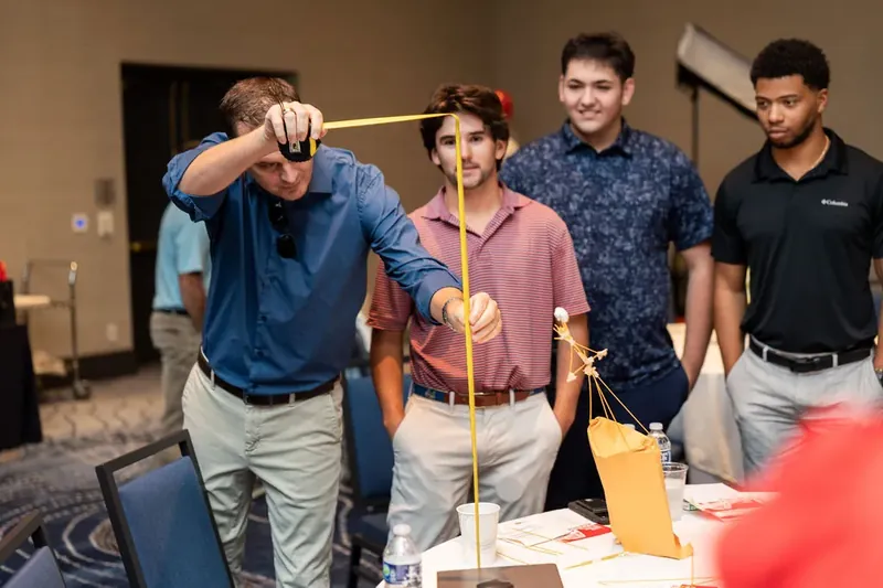 Four men work together to stabilize the wooden tower, leaning in with focused expressions as the structure grows taller.
