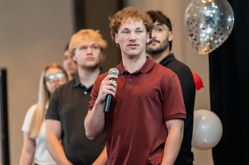 Man in burgundy polo speaking into microphone with colleagues nearby as red and silver balloons float overhead.