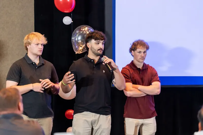 Three men in casual shirts stand together during presentation, center man holding microphone while gesturing to audience.