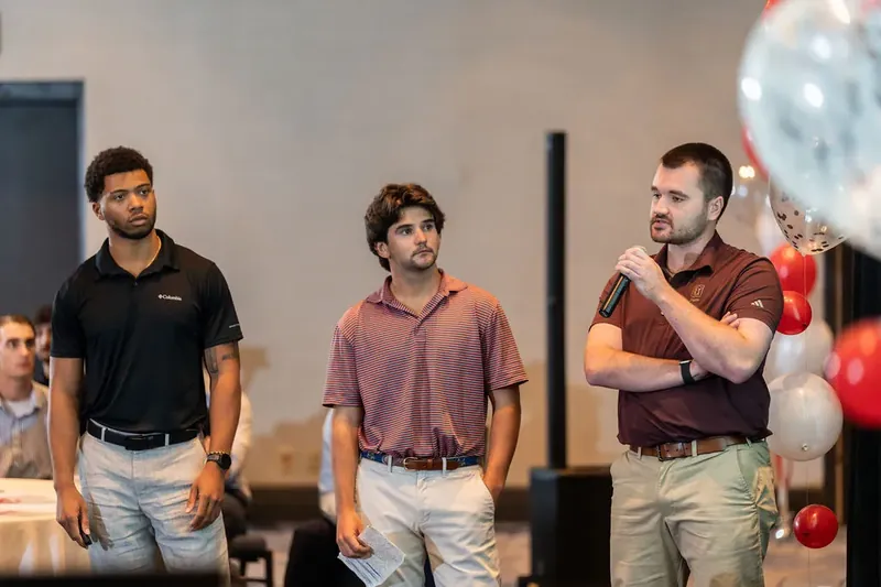 Three men in polo shirts stand side-by-side with red and white balloons suspended behind them in indoor venue.