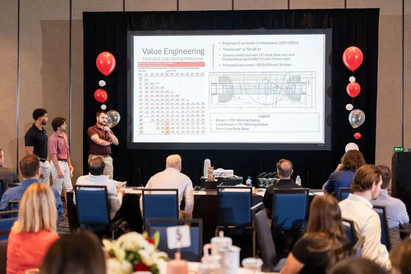 Three speakers present to seated audience with large screen displaying "Value Engineering" content and decorative balloons