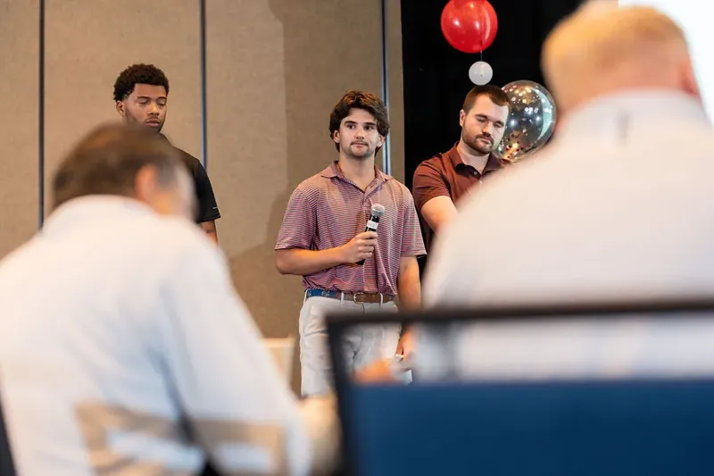 Center presenter holds microphone while speaking to audience members in foreground at corporate gathering with balloon décor.