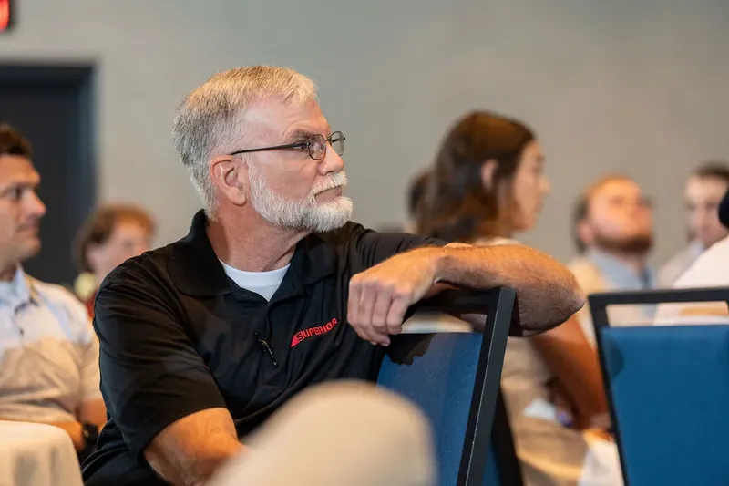 Man with gray beard and glasses in black shirt leans against chair, listening intently during indoor corporate gathering.