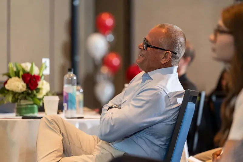 Man in light blue shirt and glasses sits relaxed at banquet table with red and white balloon décor nearby.