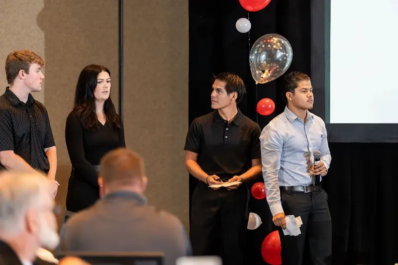 Four young professionals stand presenting together, with red and white balloons and projection screen behind them.